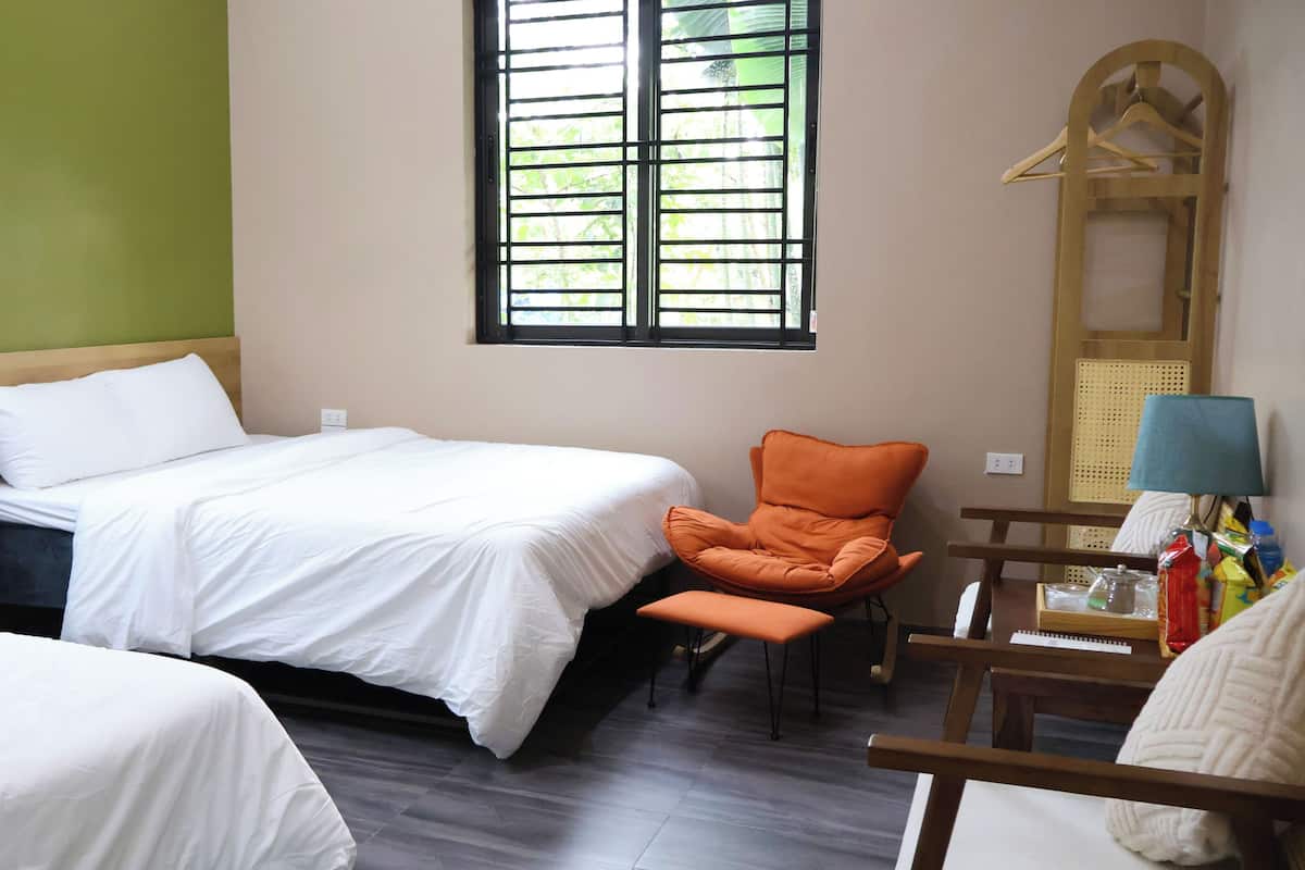 A hotel room with two beds, a wooden chair, a table with a lamp, and a window with blinds.