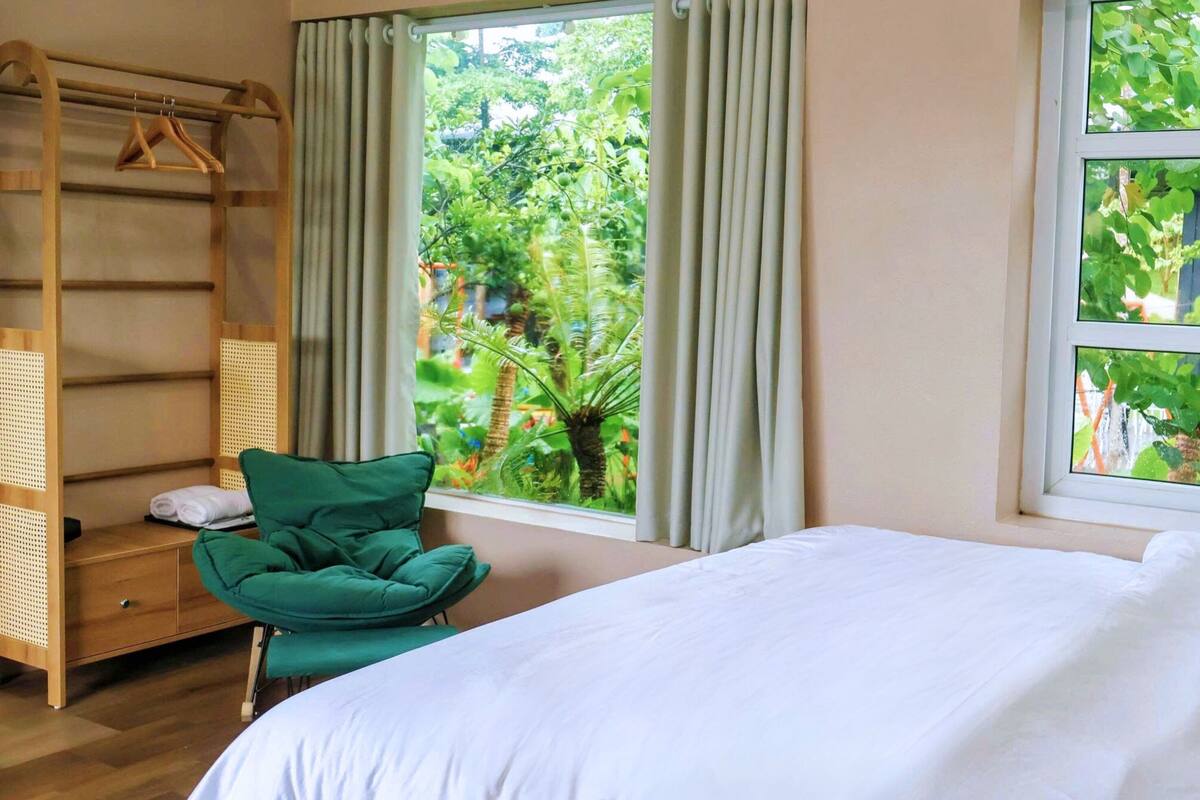 A bedroom with a bed, a green chair, a wooden shelf, and a window with a view of greenery.