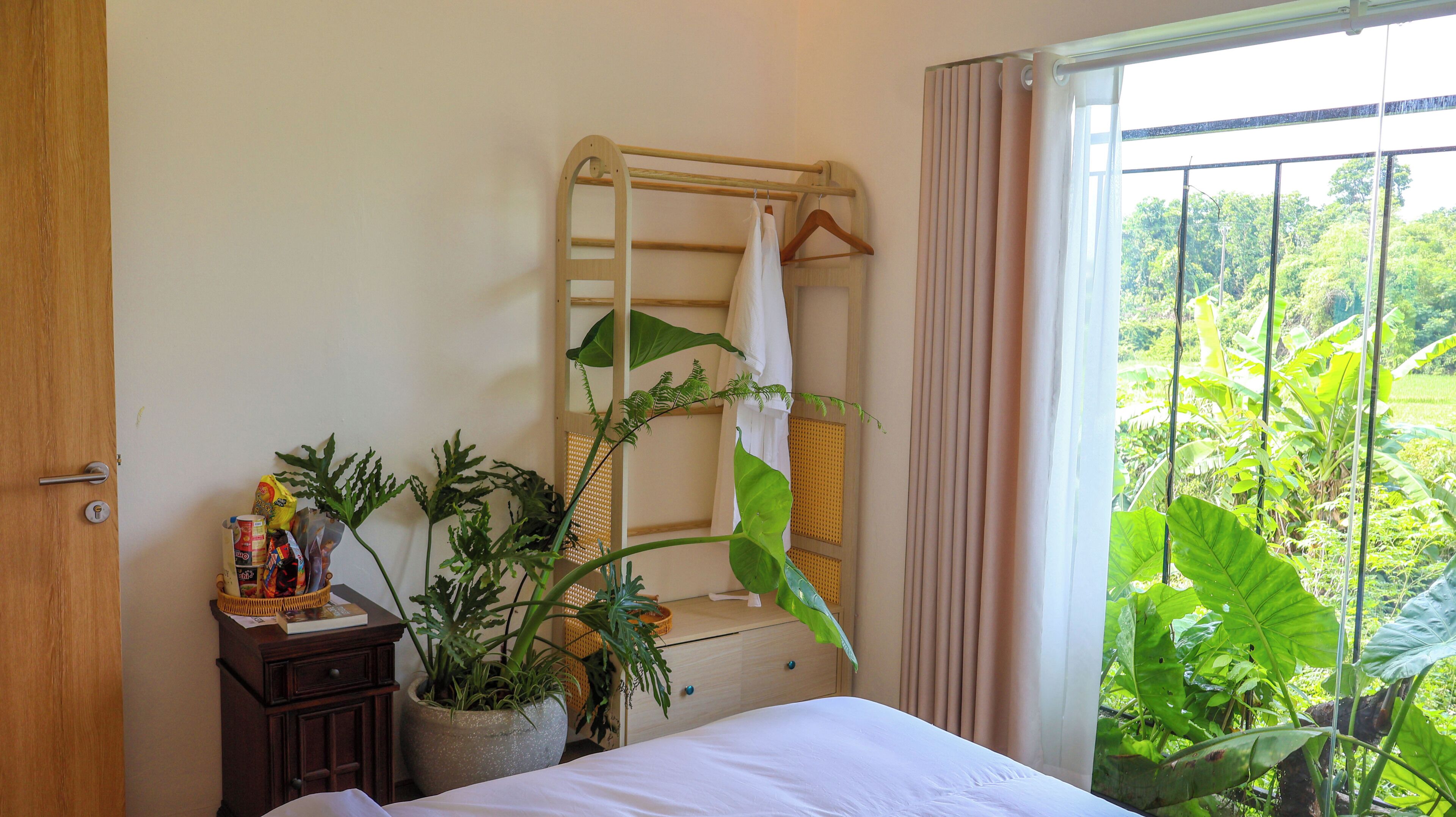 Een slaapkamer met een bed, een houten dressoir, een kamerplant, een droogrek en uitzicht op groen door een raam.