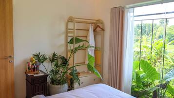 Een slaapkamer met een bed, een houten dressoir, een kamerplant, een droogrek en uitzicht op groen door een raam.