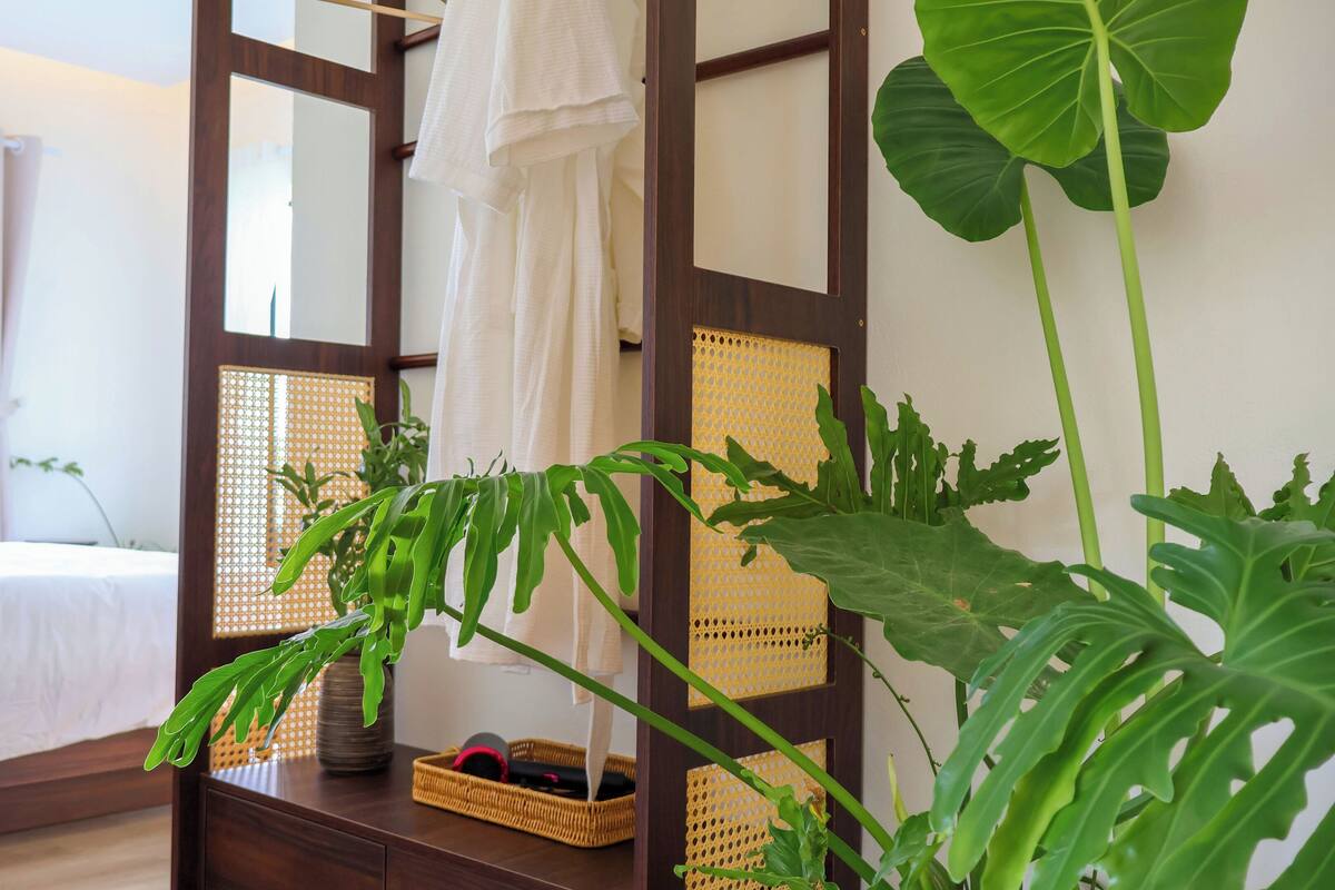 A hotel room with a bed, a wooden side table with a basket, and large green plants.
