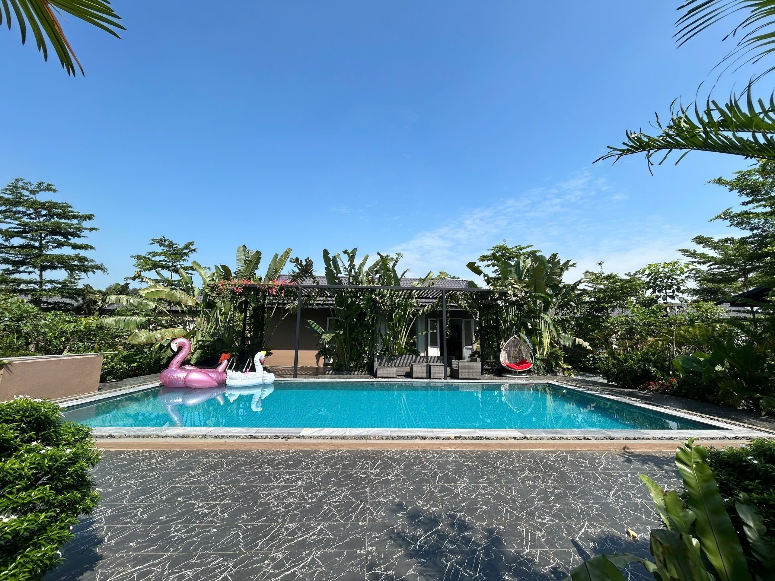 A swimming pool with a pink flamingo float and a swan float, surrounded by lush greenery and a covered seating area.