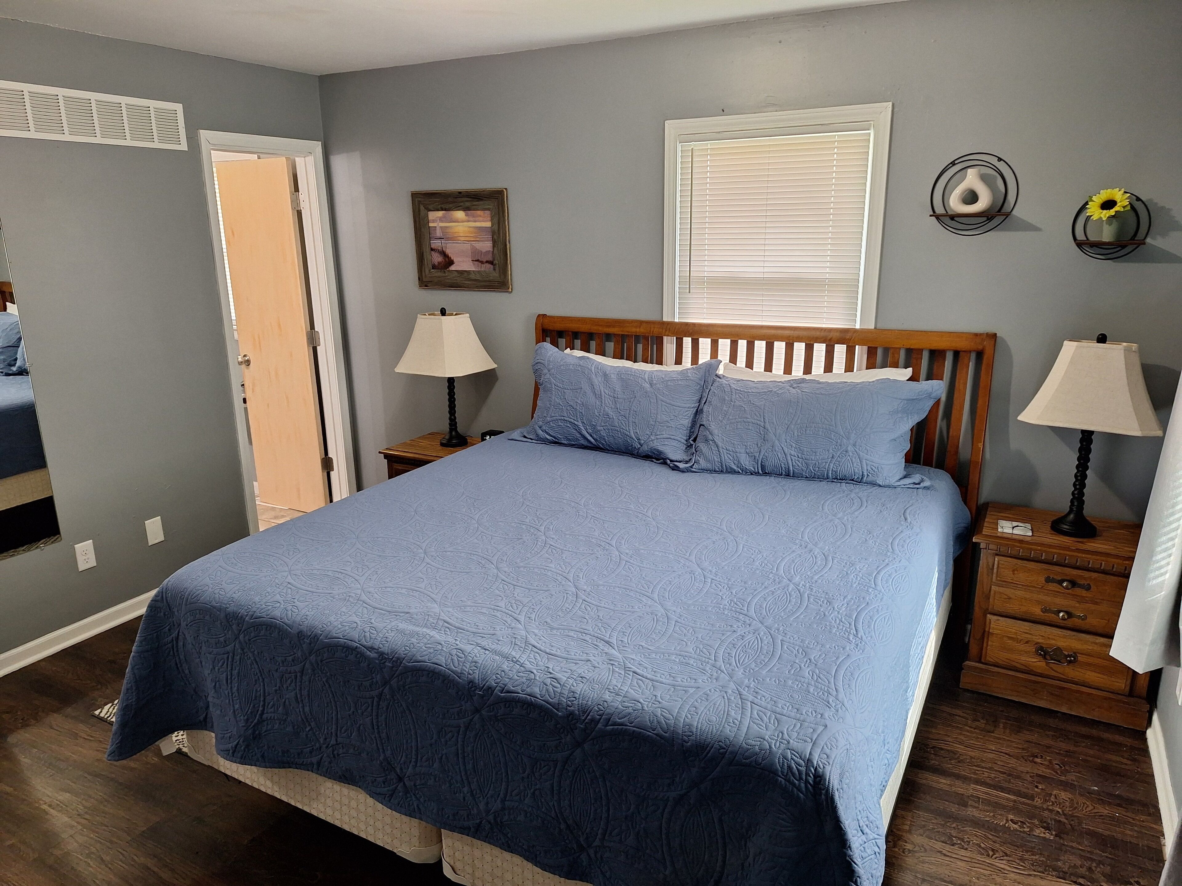 A bedroom with a large bed, two bedside tables with lamps, a window with blinds, a door, and a wall-mounted shelf with decorative items.