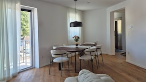 Una sala de estar moderna con un sofá, una mesa de comedor y sillas. Hay un ventanal con cortinas, una lámpara colgante y una puerta que da a otra habitación.