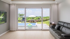 A spacious living room with a large sliding glass door leading to a pool area, a black leather sofa, and a flat-screen TV mounted on the wall. - Gimnyeong stonewall Private Pool Villa (Jeju City)