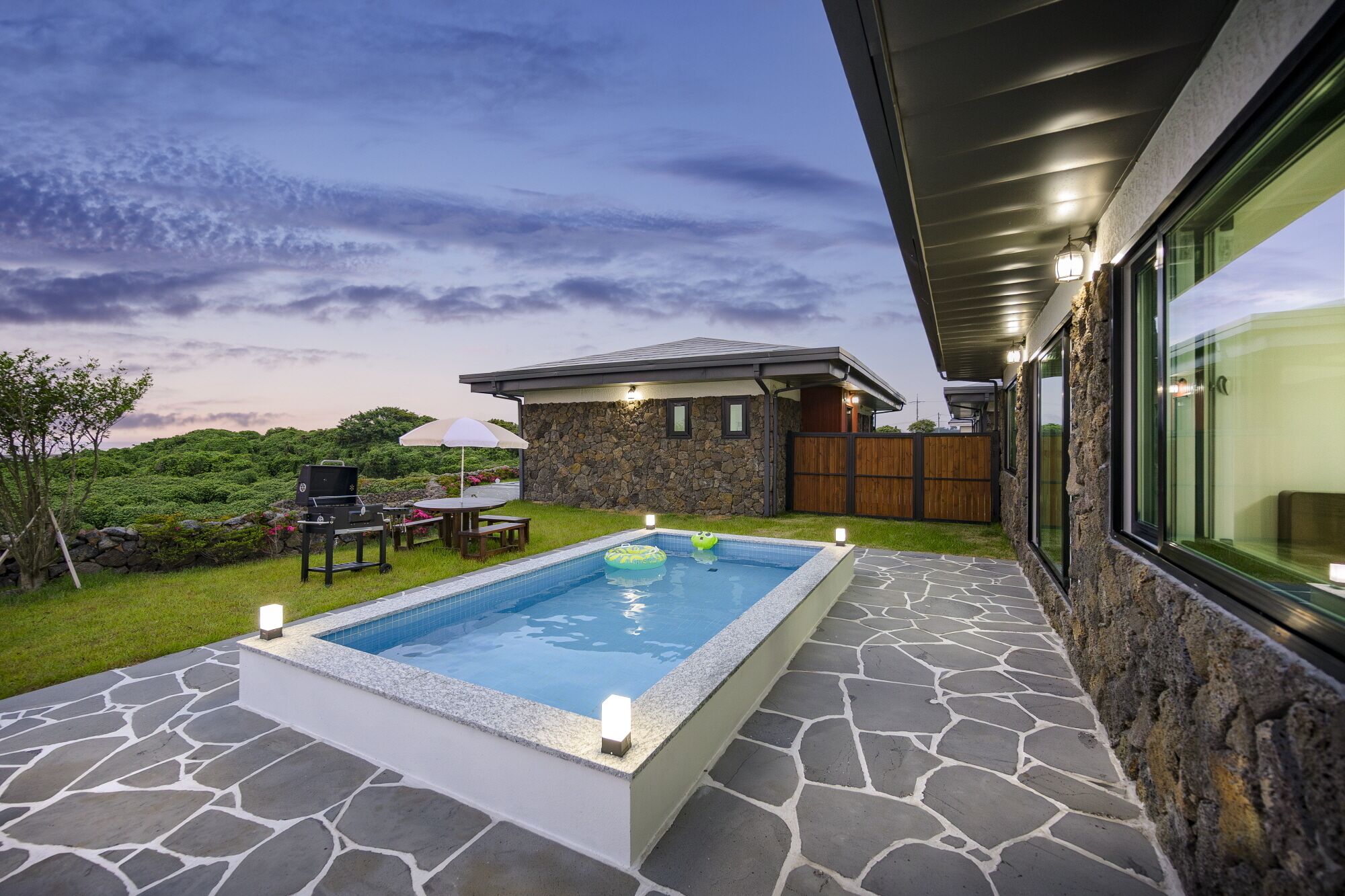 A swimming pool with stone tiles, a barbecue area, and a stone house with a wooden fence.