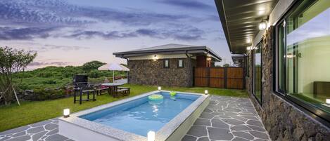 A swimming pool with stone tiles, a barbecue area, and a stone house with a wooden fence.
