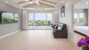A spacious living room with a large TV, a grey sofa, and a view of a pool and greenery. - Gimnyeong stonewall Private Pool Villa (Jeju City)