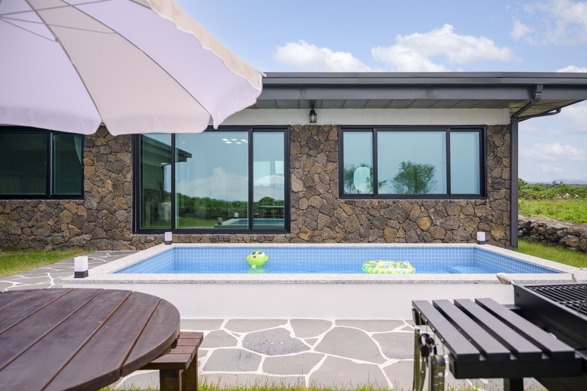 Een modern huis met een gevel van natuursteen, grote ramen, een zwembad en tuinmeubilair.
