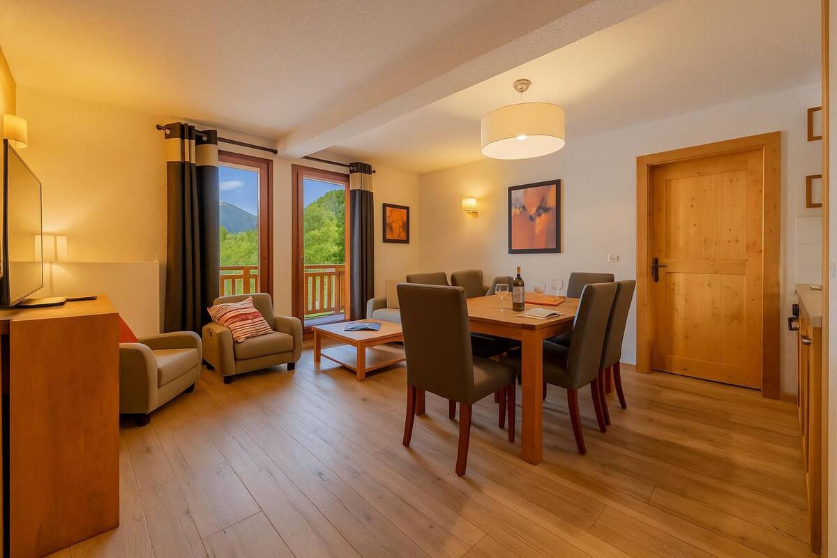 A room with a wooden dining table, chairs, a sofa, a TV, and a window with curtains.