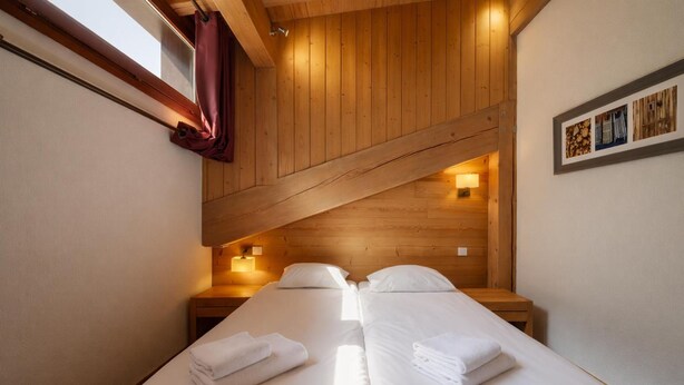 A room with two beds, wooden ceiling beams, a window with curtains, and wall-mounted lights. - l Ours Bleu Apartments (Vallorcine)