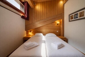 A room with two beds, wooden ceiling beams, a window with curtains, and wall-mounted lights. - l Ours Bleu Apartments (Vallorcine)