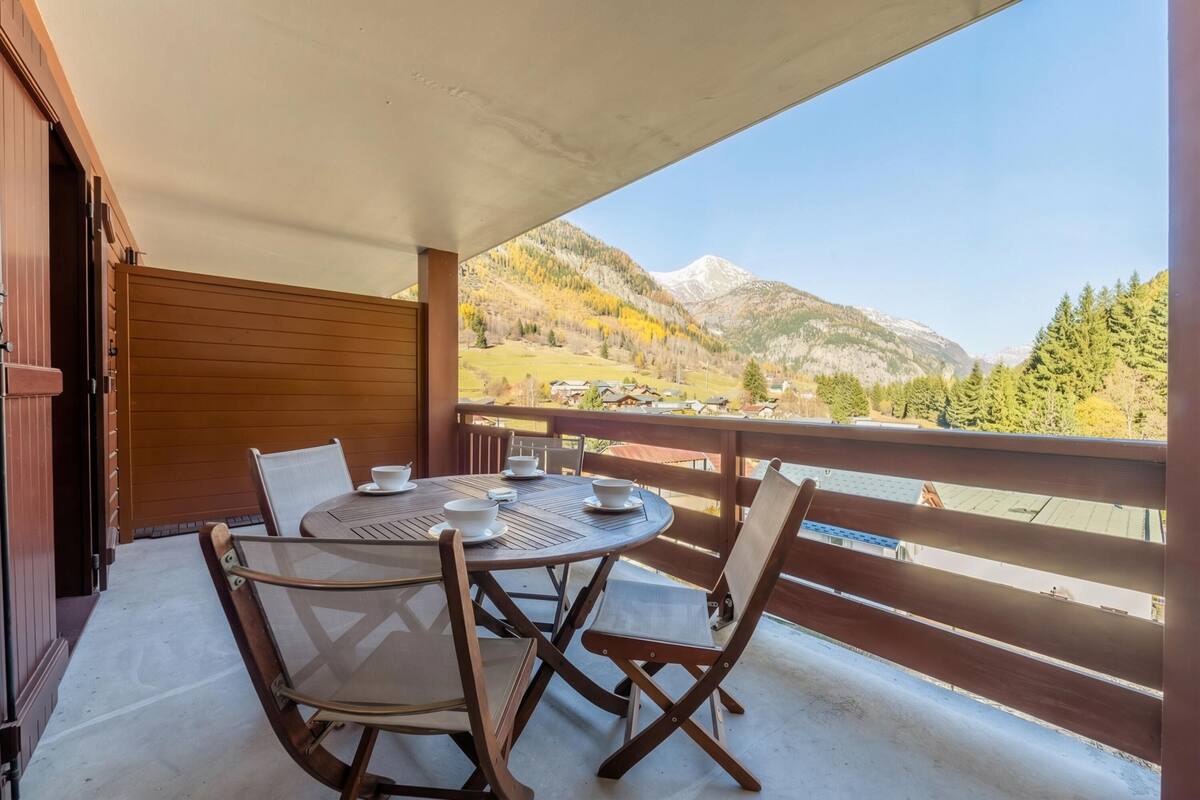 A balcony with a round table and chairs, a wooden railing, and a mountain view.