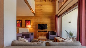 A cozy living room with a wooden ceiling, a large window with curtains, a sofa, two armchairs, a coffee table, and a flat-screen TV on a wooden stand. - l Ours Bleu Apartments (Vallorcine)
