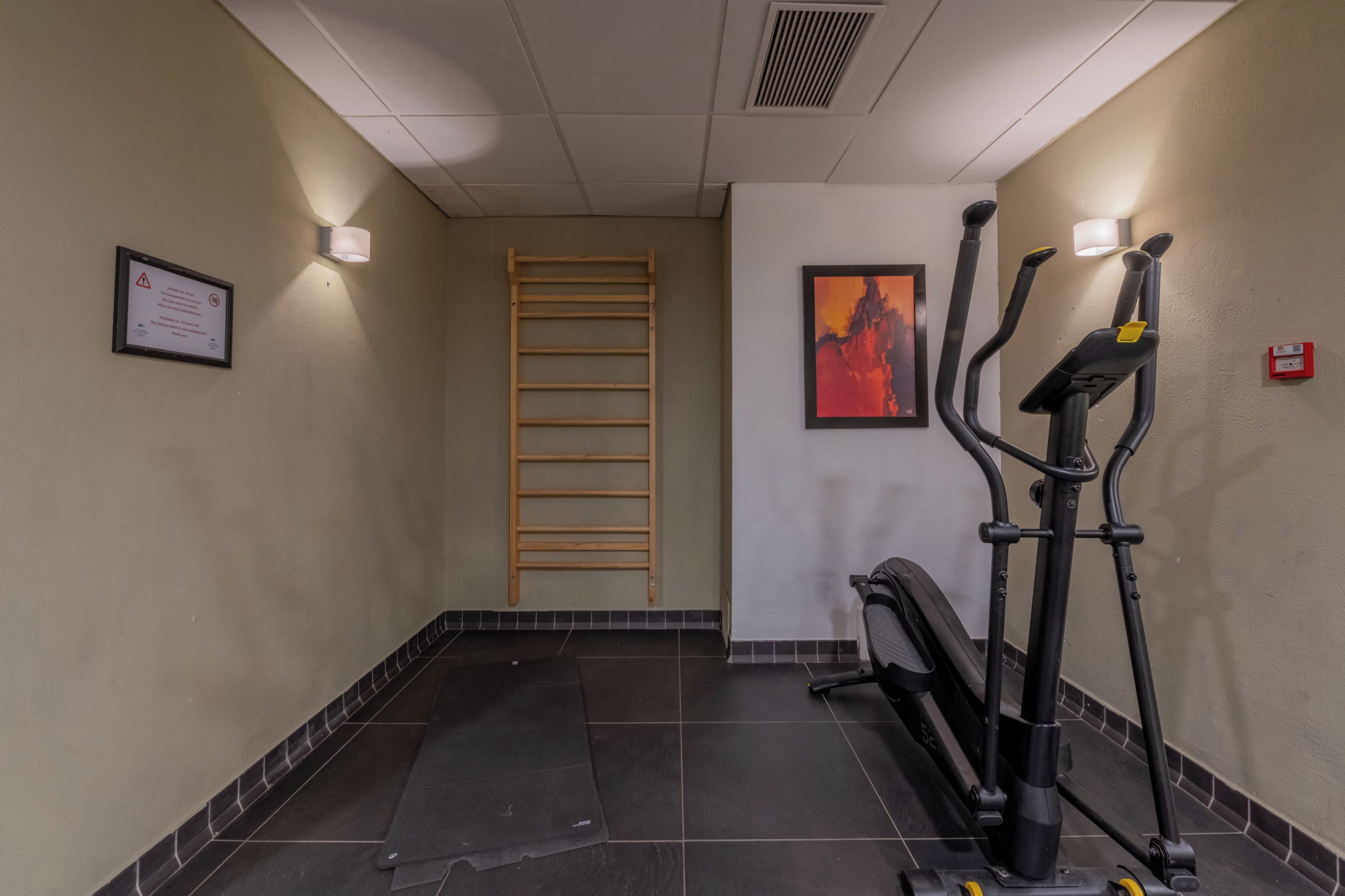 A hallway with a treadmill, a wall-mounted light fixture, a framed notice, and a wooden ladder.