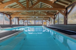 Une piscine intérieure avec des parois en verre transparent, des poutres en bois et un sol carrelé.