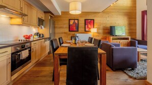 A modern kitchen with wooden cabinets, a dining table with chairs, and a TV in the background. - l Ours Bleu Apartments (Vallorcine)