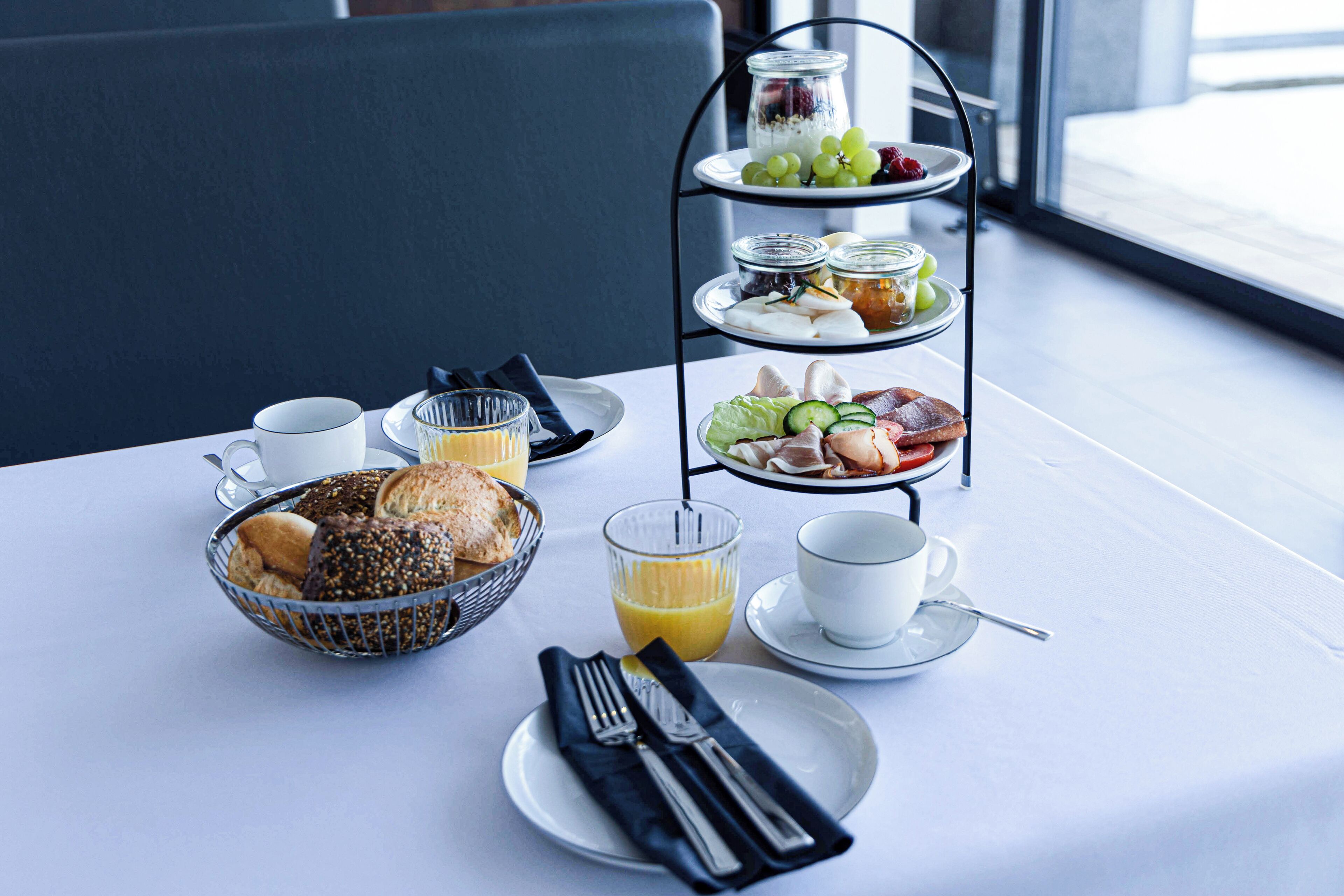A breakfast setup with a three-tiered tray, a bowl of pastries, a glass of orange juice, and a cup of coffee on a table with a view of the outside.