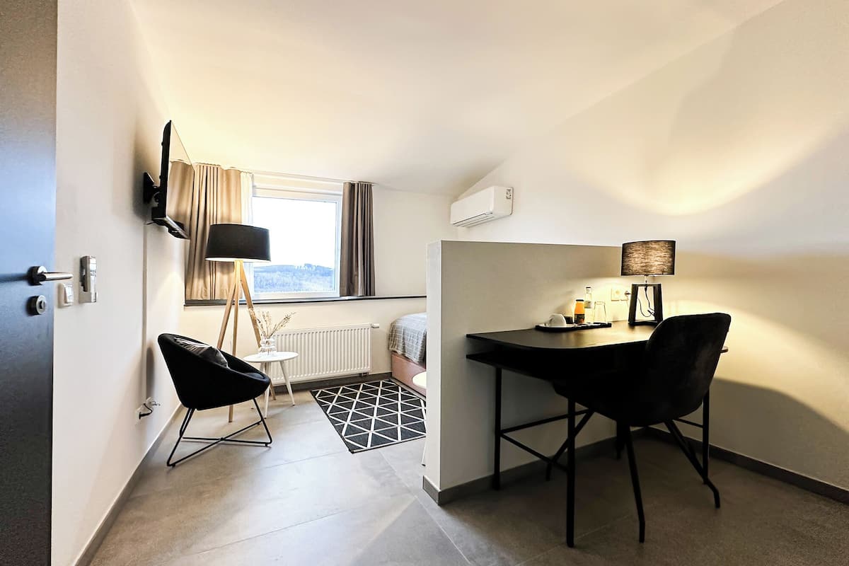 A compact hotel room with a bed, a small table with a chair, a floor lamp, and a radiator.