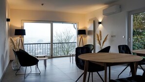 Een moderne woonkamer met een grote schuifdeur naar een balkon, een ronde houten tafel en twee zwarte stoelen.