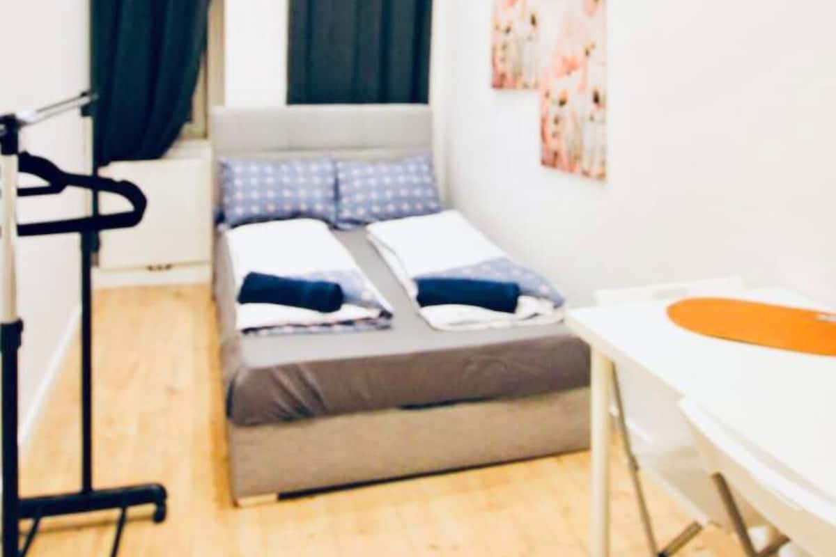 A small, narrow room with a bed, a desk, and a chair. There are two windows with dark curtains, and the walls are white.