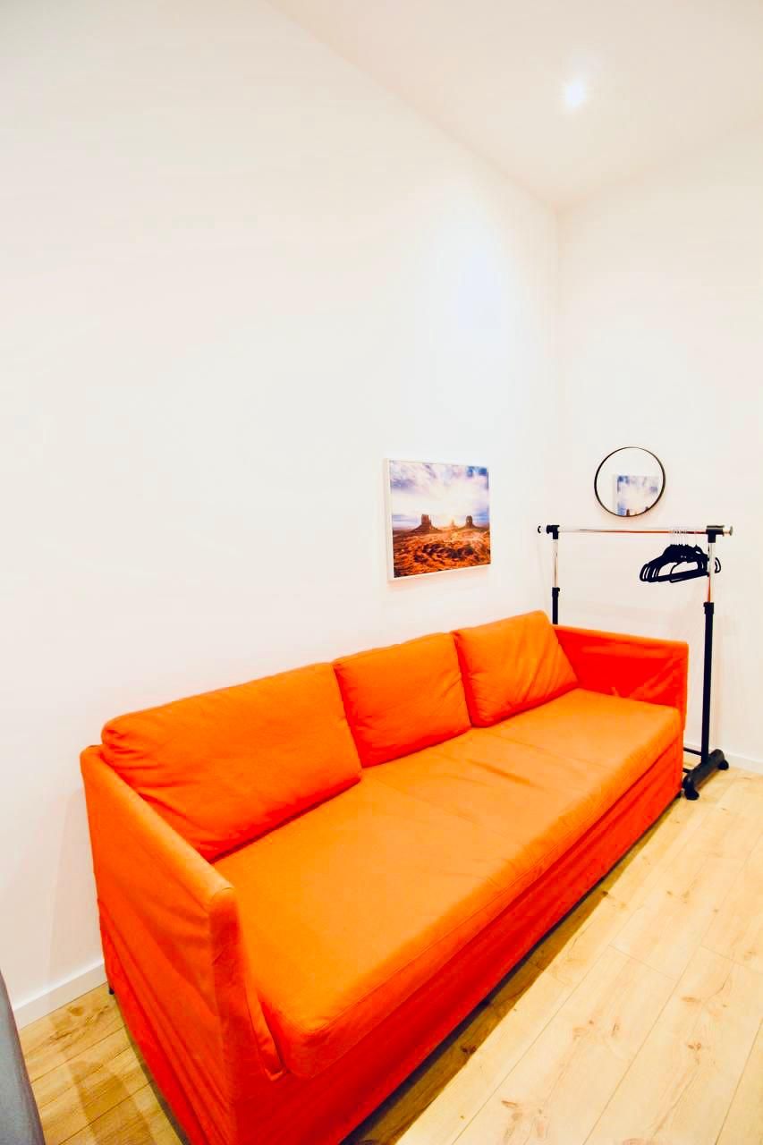 Ein Zimmer mit einem leuchtend orangefarbenen Sofa, einem kleinen gerahmten Bild an der Wand und einem Schuhregal mit Spiegel.