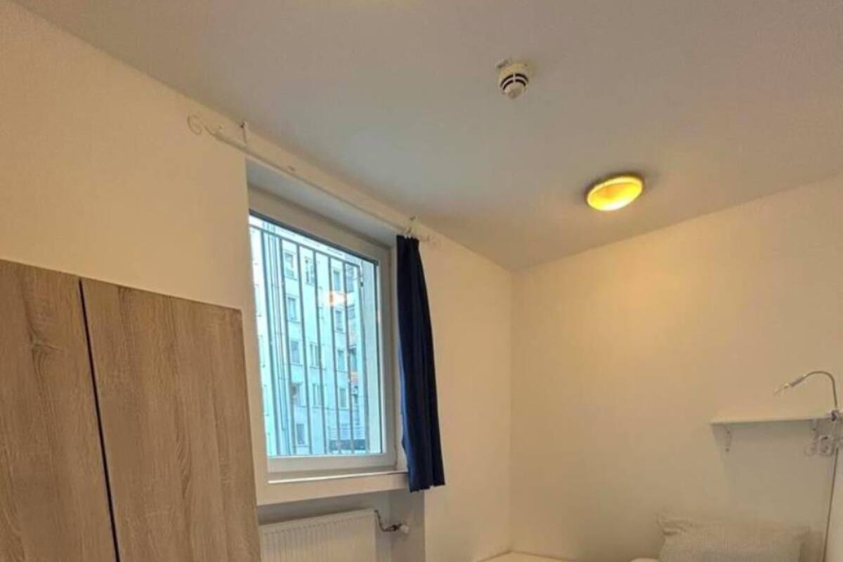 Une chambre avec une armoire en bois, une petite table de chevet, un lit avec une literie blanche, un radiateur et une fenêtre donnant sur des immeubles.
