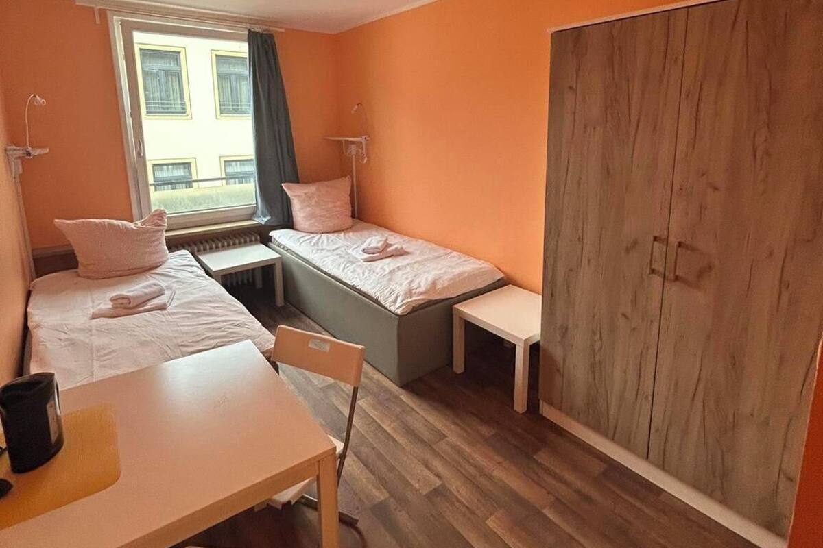 Une chambre d’hôtel compacte comprenant deux lits simples, une armoire en bois, une petite table avec une chaise et une fenêtre avec des rideaux.