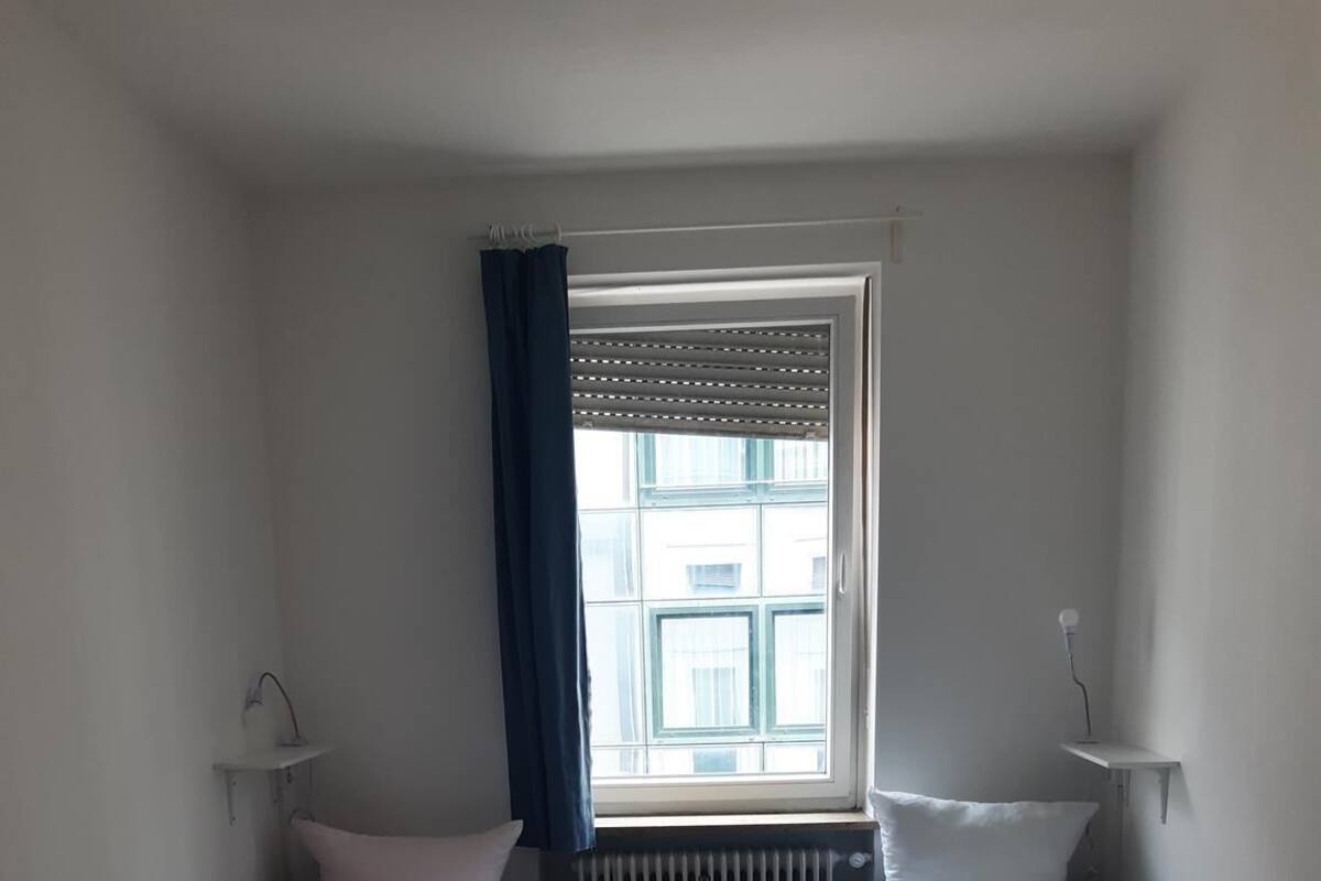 Une petite chambre d’hôtel composée d’une seule pièce, avec deux lits simples, une fenêtre avec des stores, un radiateur et une petite table.