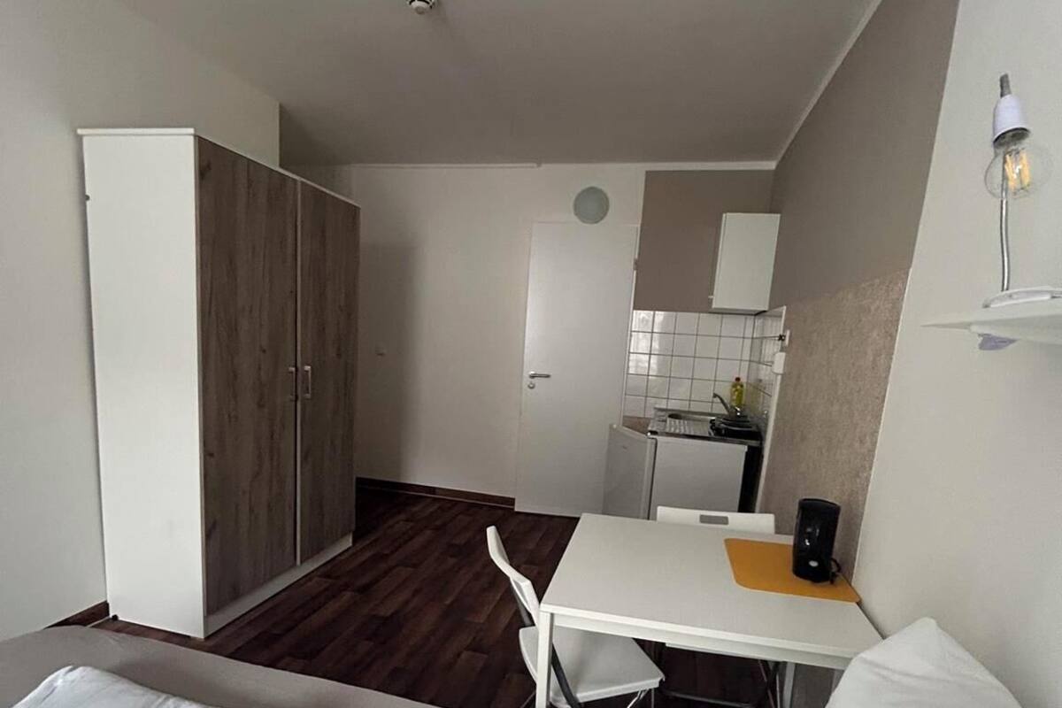 Une pièce de taille modeste comprenant un lit, une petite table, une chaise, une armoire et un coin cuisine.