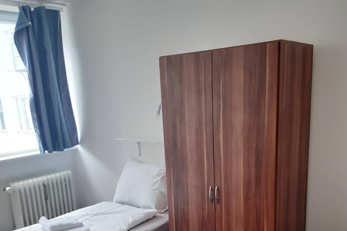 Une chambre avec une armoire en bois, un lit avec une literie blanche, un radiateur et une fenêtre avec des rideaux bleus.