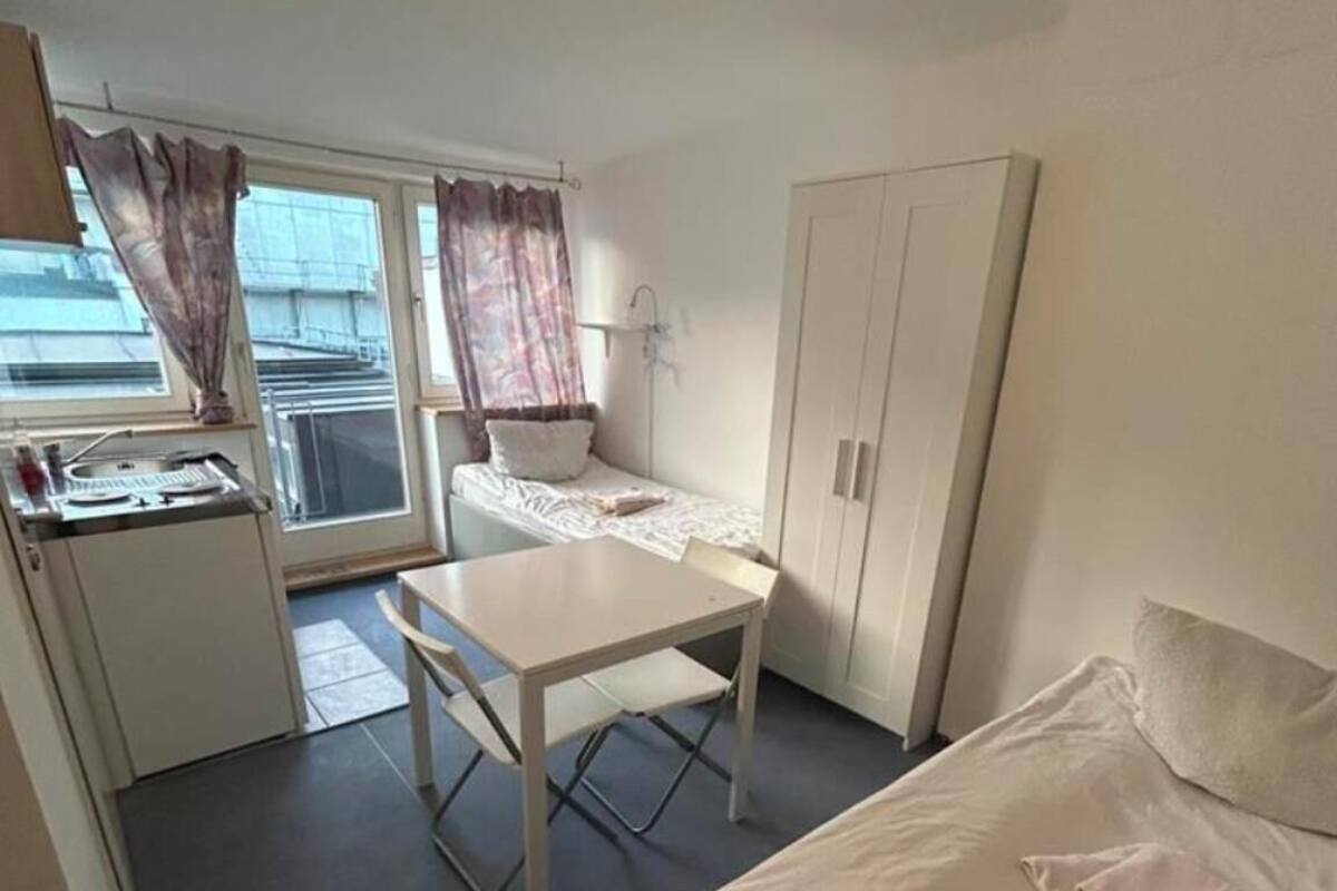 Une petite pièce, de taille modeste, comprenant un lit, un bureau et une armoire. Elle est dotée d’une fenêtre avec des rideaux, d’un lavabo et d’une porte donnant sur un espace extérieur.
