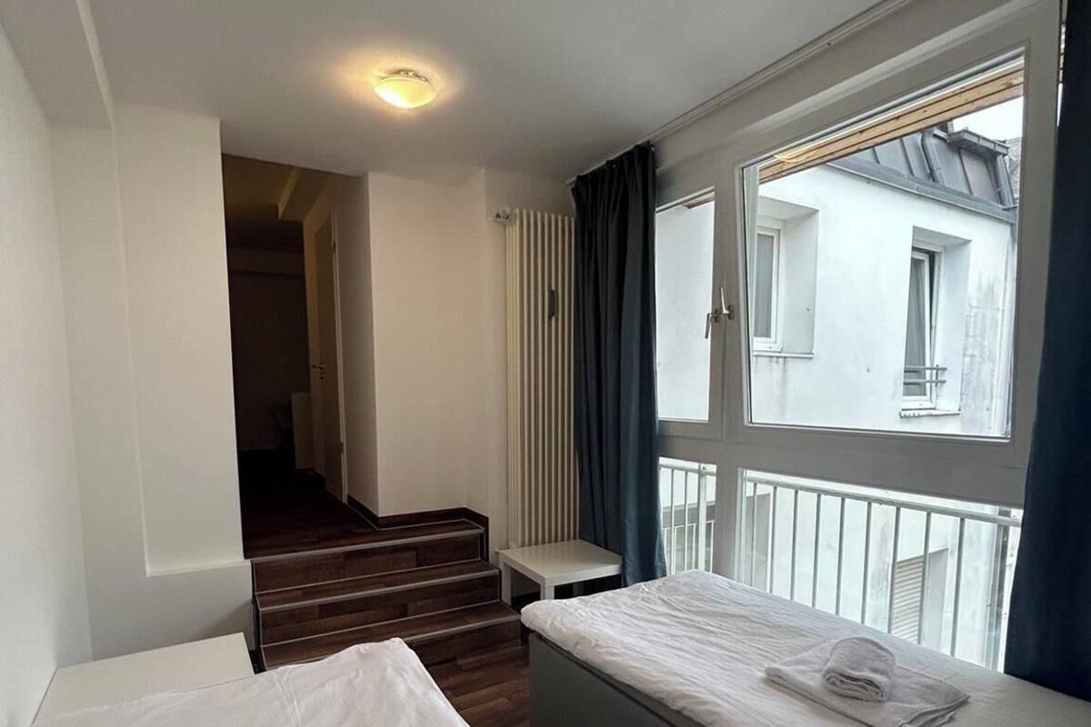 Une chambre d’hôtel avec deux lits, une fenêtre avec des rideaux, un radiateur et un escalier menant à une autre pièce.