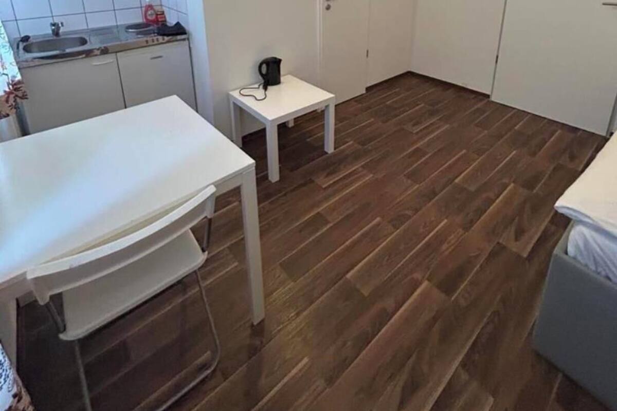 Une petite pièce propre, avec un plancher en bois, une table blanche, un petit meuble blanc et un lit blanc.