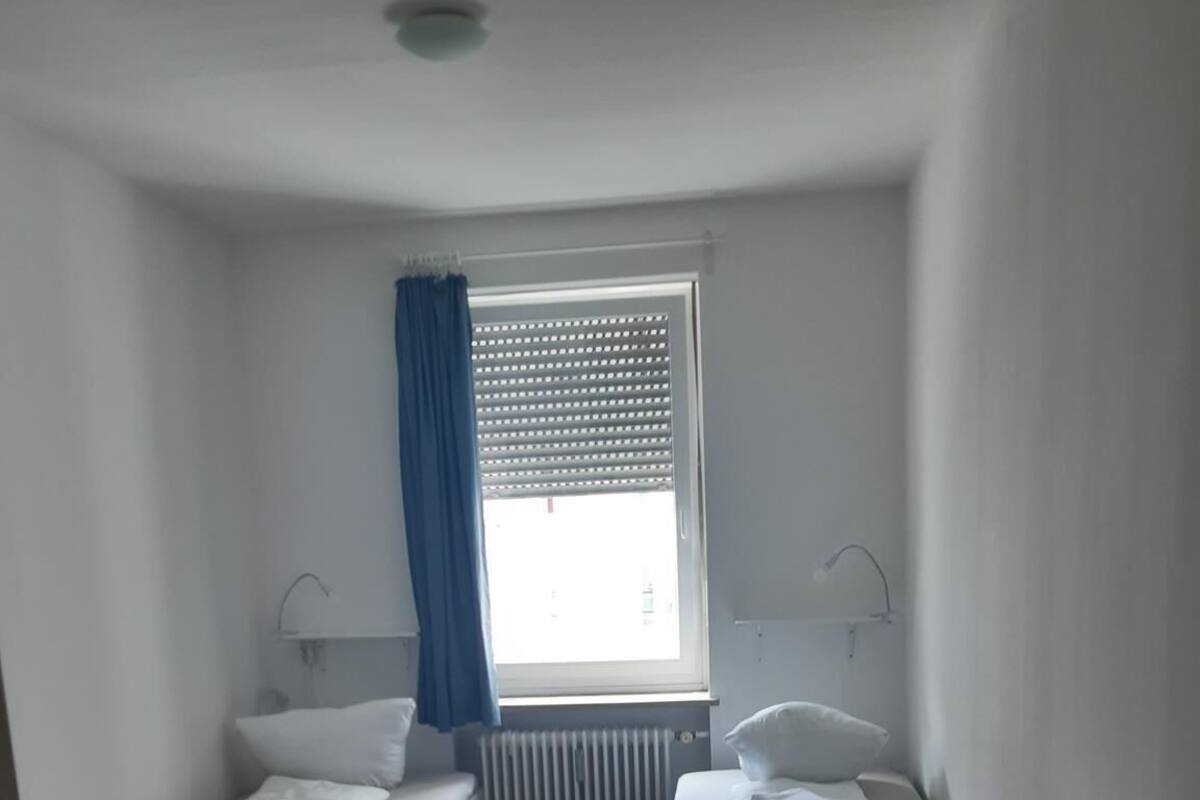 Une petite pièce bien éclairée, avec un lit, un bureau et une chaise. On y trouve une fenêtre avec des stores, un radiateur et un rideau.