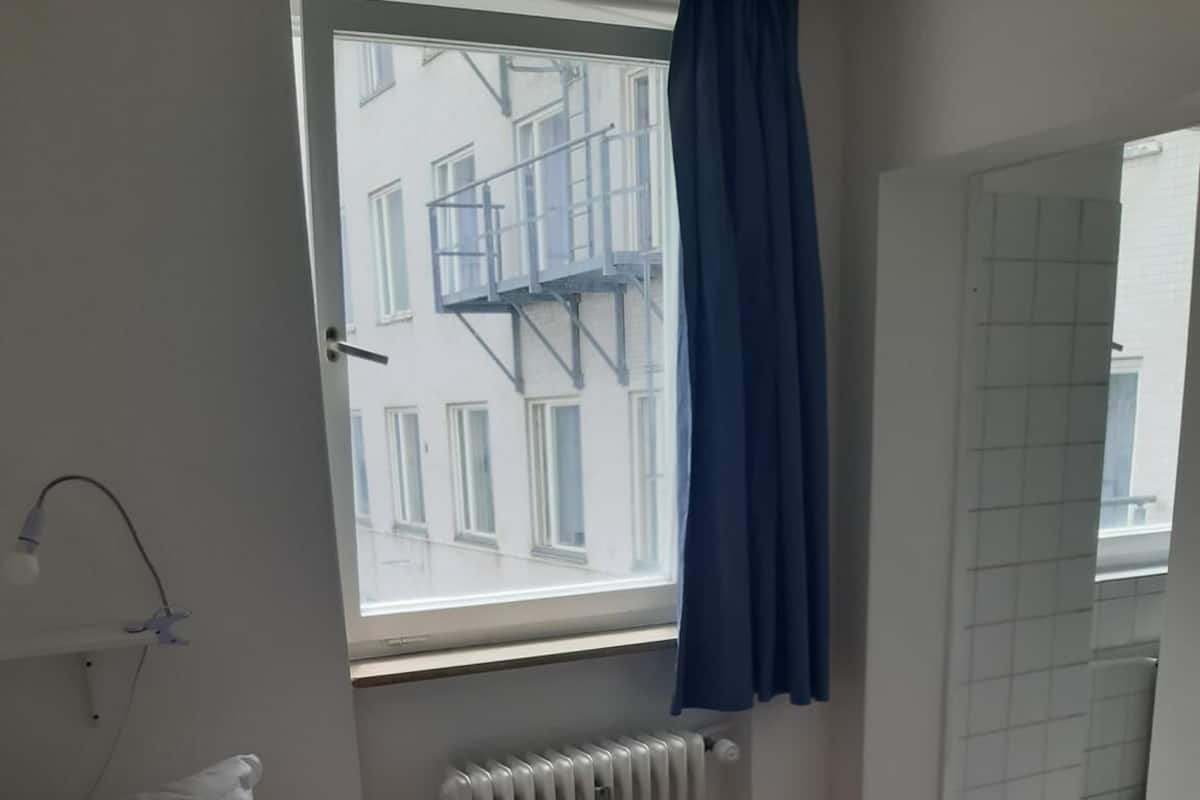 Une chambre à coucher avec un lit, une fenêtre donnant sur des immeubles, un radiateur et une table.
