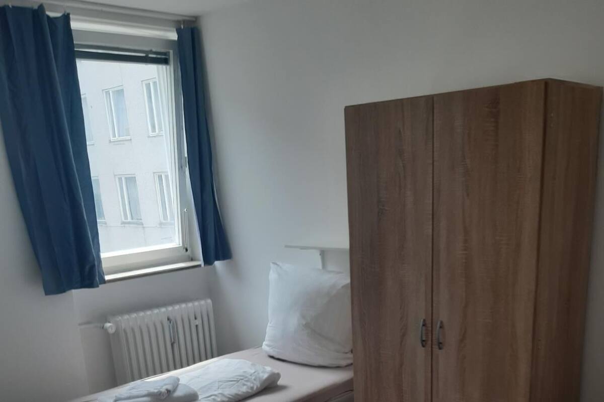 Une petite chambre à coucher, bien rangée, avec un lit simple, une armoire en bois et une fenêtre avec des rideaux.