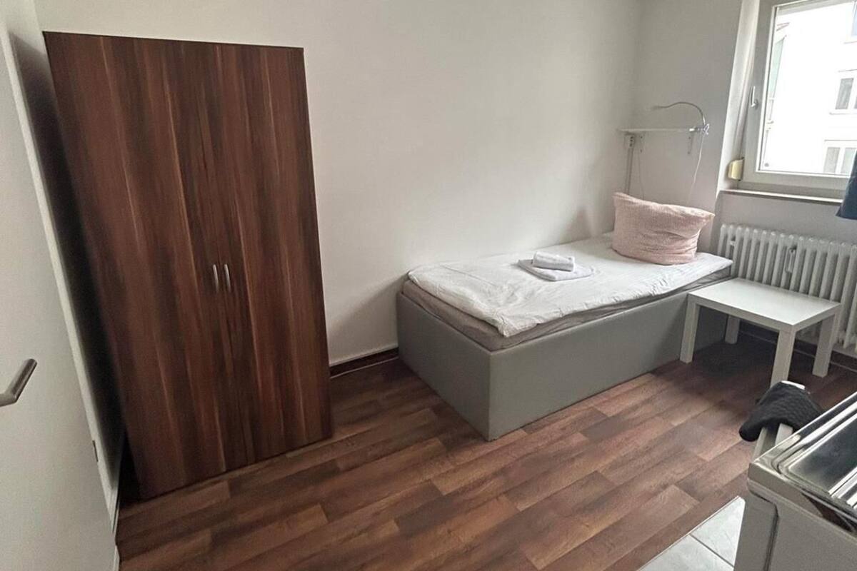 Une petite pièce, de taille modeste, comprenant une armoire en bois, un lit avec un matelas et des oreillers, une petite table et un radiateur.
