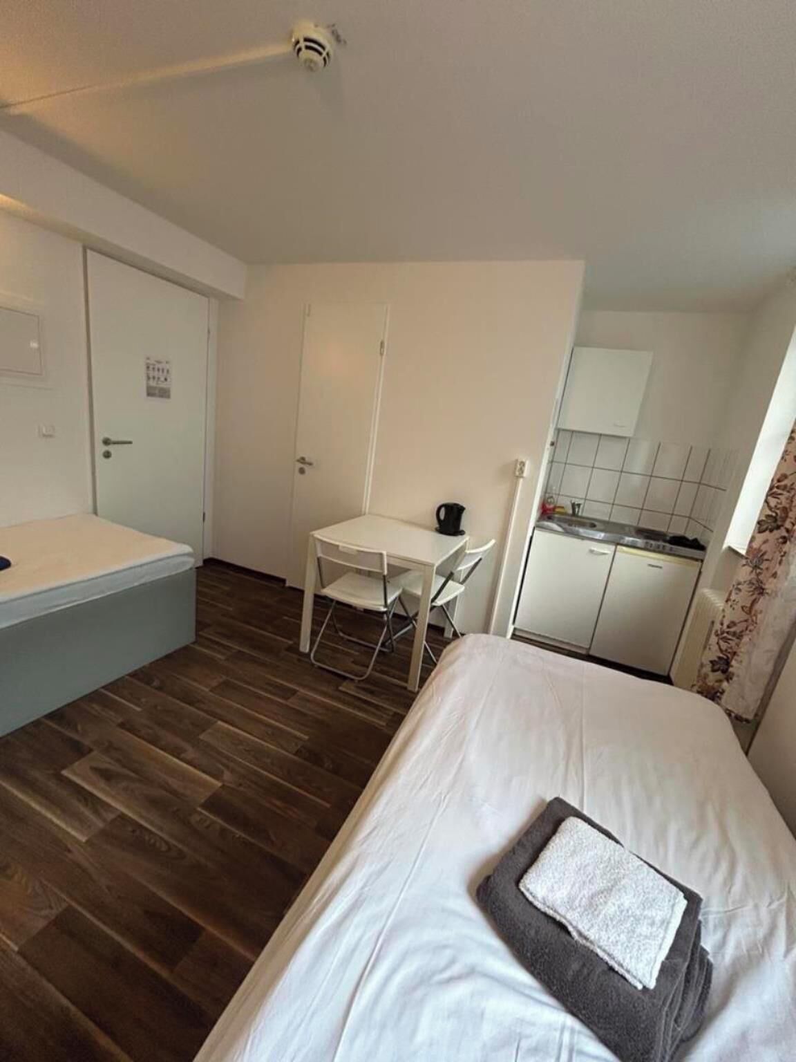 Ein kleines Hotelzimmer mit einem Bett, einem Schreibtisch, einem Stuhl und einer Küchenzeile.