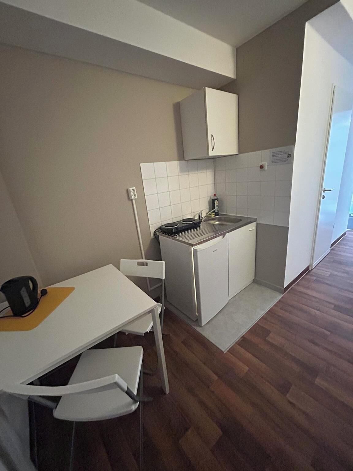 Cocina compacta con armarios blancos, una pequeña encimera y un lavamanos. Hay una mesa de comedor con sillas y una tetera encima.