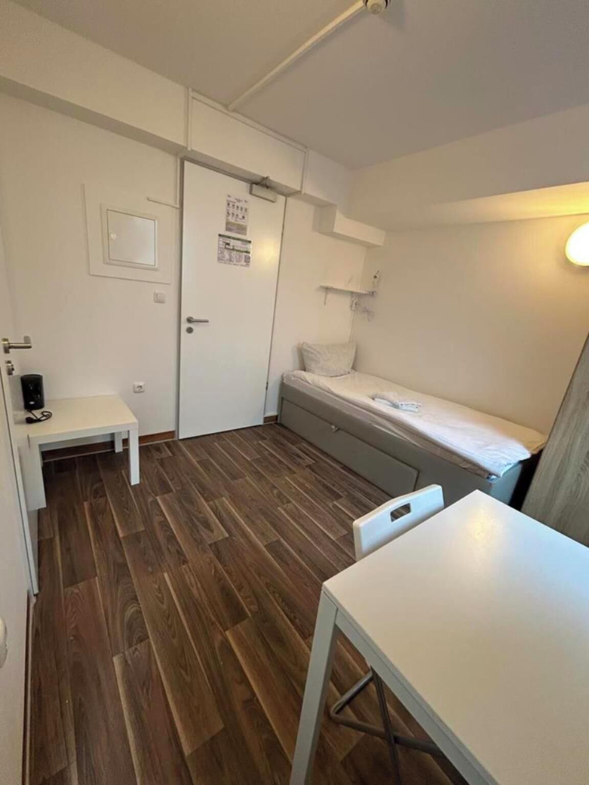 A small, compact room with a bed, a desk, and a chair. There is a door with a poster on it, and a small table with a lamp.
