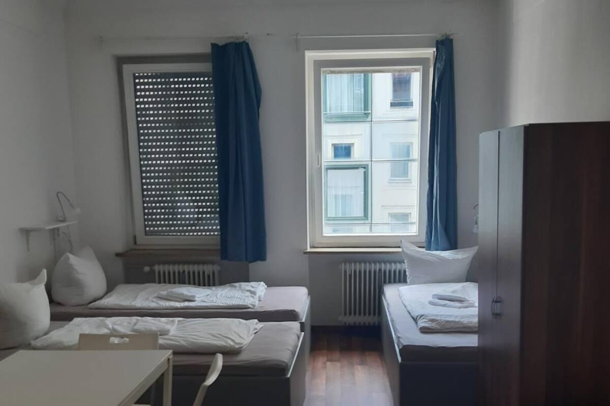 Une petite pièce bien éclairée, avec deux lits, une table et une chaise. Il y a une fenêtre avec des rideaux bleus, d’où l’on peut voir les bâtiments à l’extérieur.
