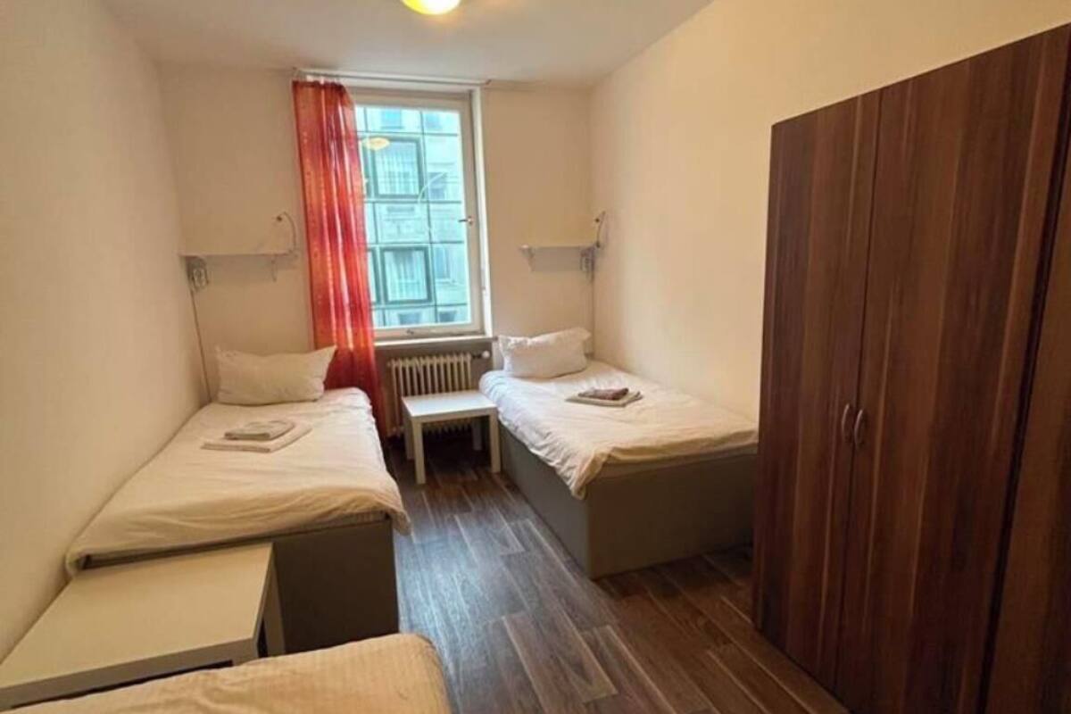 Une petite chambre, de taille modeste, comprenant deux lits simples, une armoire en bois, une fenêtre avec des rideaux et un radiateur.