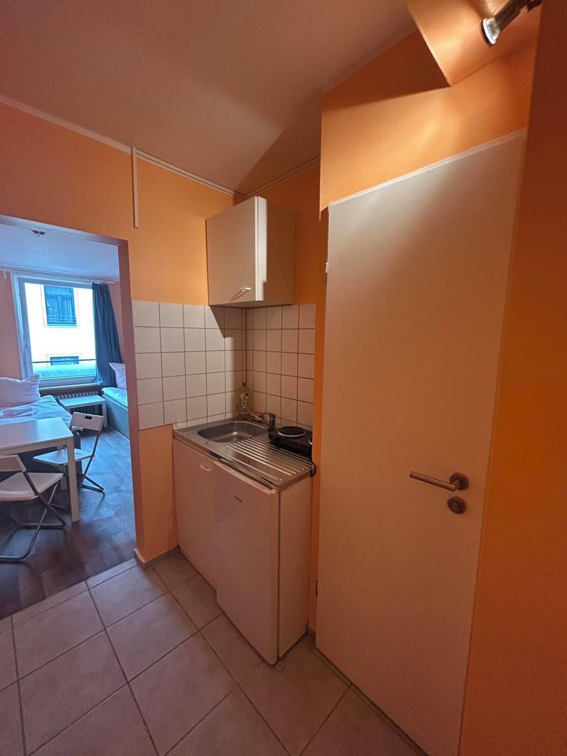 A compact kitchen with a sink, a stove, and a cabinet. There is a dining area with a table and chairs visible through an open door.