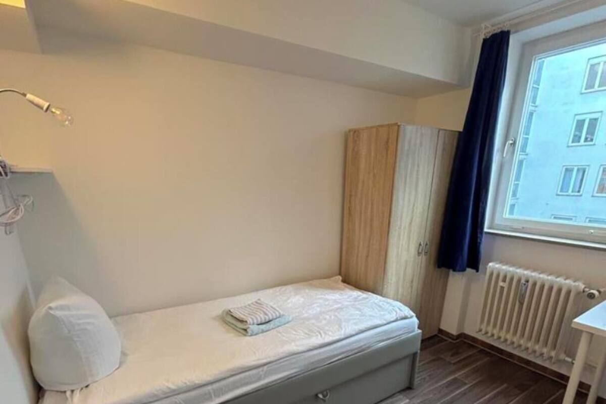 Une petite chambre avec un lit simple, un plancher en bois, une fenêtre avec des rideaux, un radiateur et une petite table.