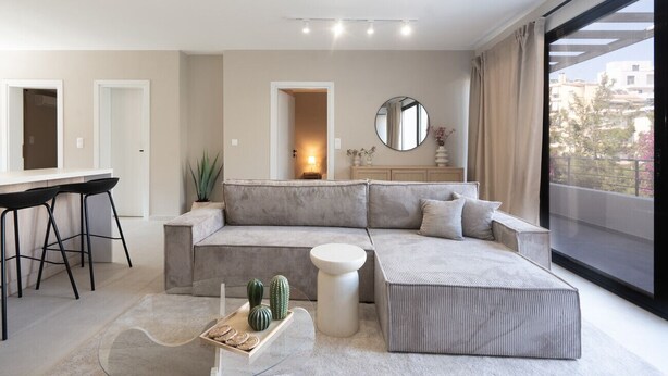 A modern living room with a sectional sofa, a dining area with bar stools, a round mirror, and a large window with a view of buildings. - LIV - Deluxe 2BD Apartment in Athens (Athens)