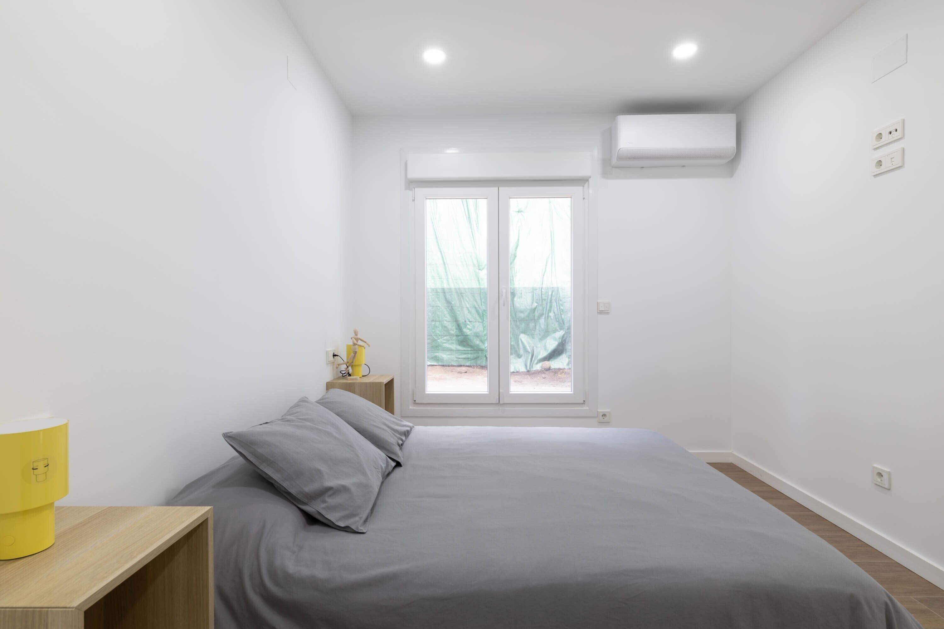 Un dormitorio con cama, mesita de noche, ventana y equipo de aire acondicionado.