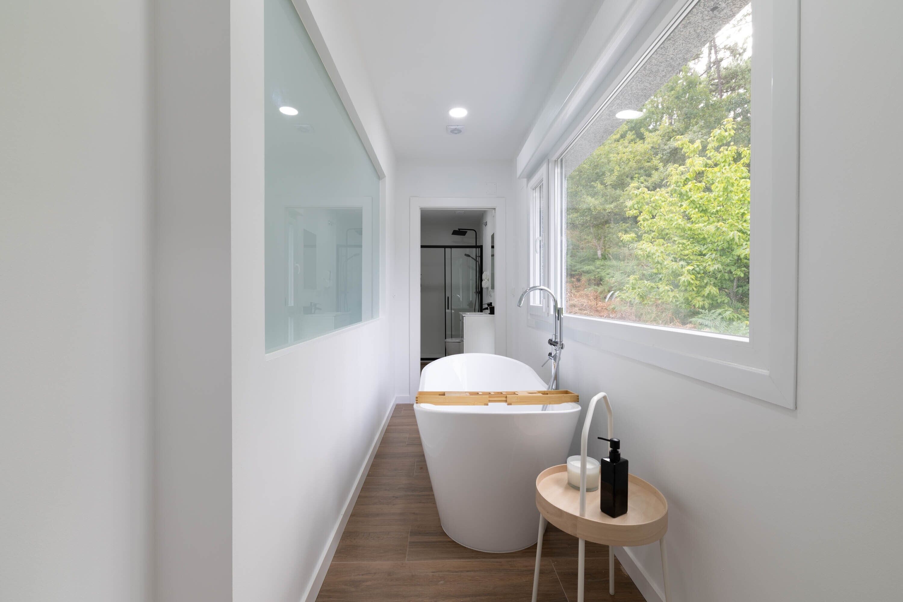 Un baño moderno con una bañera independiente, un taburete de madera pequeño y un ventanal con vistas a la vegetación.