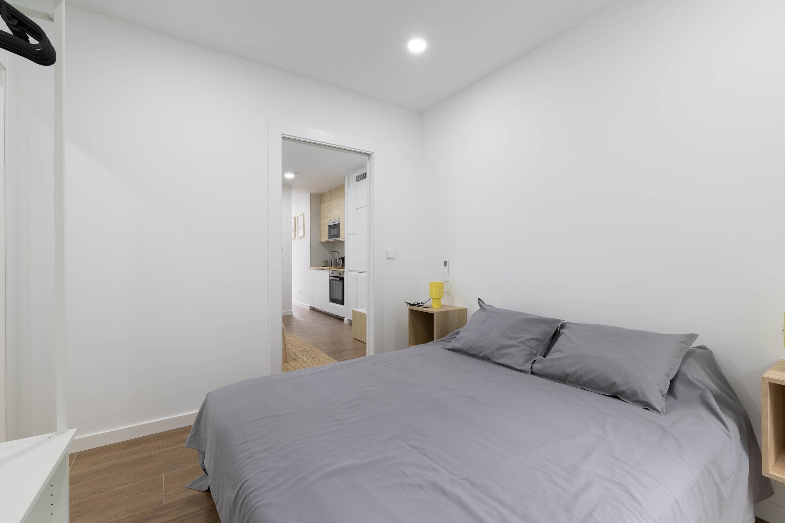Un dormitorio con cama, mesita de noche y una pequeña cocina visible a través de una puerta abierta.
