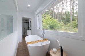 A modern bathroom with a freestanding bathtub, a glass shower enclosure, and a view of a forest through a large window. - Candida Apartment with Mountain Views, Shared Garden, and Air Conditioning (San Cosme-Outes)